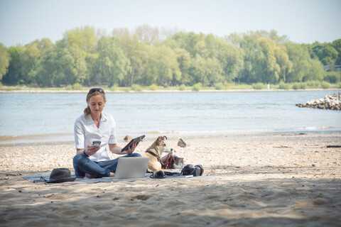 Woman sitting on blanket at a river with dog using portable devices stock photo