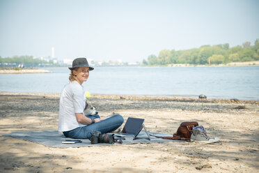 Smiling woman sitting on blanket at a river with dog and laptop - ONF01131