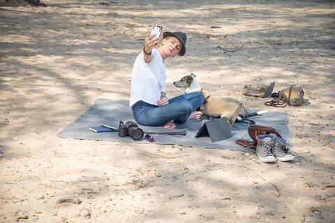 Frau sitzt auf einer Decke am Strand mit Hund und macht ein Selfie, lizenzfreies Stockfoto