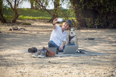 Lächelnde Frau sitzt auf einer Decke am Strand mit Hund und macht ein Selfie - ONF01133