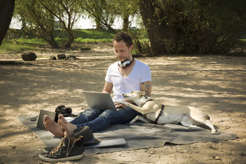 Man with dog sitting on blanket at a beach using laptop stock photo