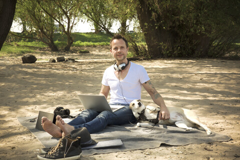 Porträt eines Mannes mit Hund, der auf einer Decke am Strand sitzt und einen Laptop benutzt, lizenzfreies Stockfoto