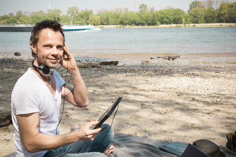 Portrait of smiling man sitting on blanket at a river using tablet stock photo