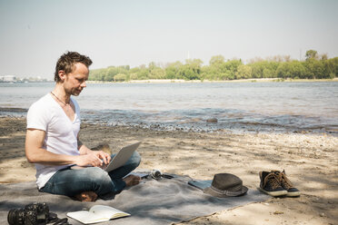 Man sitting on blanket at a river using laptop - ONF01156