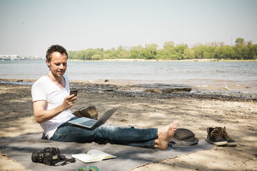 Man sitting on blanket at a river using laptop and cell phone - ONF01157