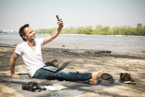 Lächelnder Mann sitzt auf einer Decke an einem Fluss und macht ein Selfie, lizenzfreies Stockfoto