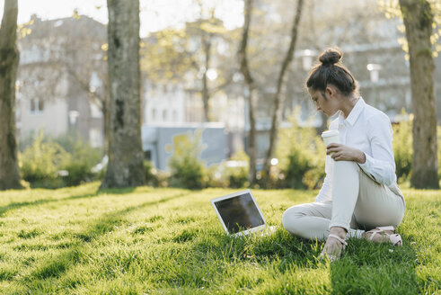 Junge Geschäftsfrau macht Pause, benutzt einen Laptop, sitzt im Gras und trinkt Kaffee - KNSF03926