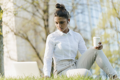 Junge Geschäftsfrau macht Pause, benutzt einen Laptop, sitzt im Gras und trinkt Kaffee - KNSF03928