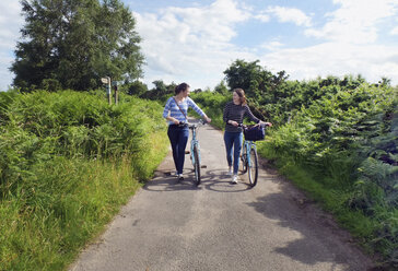 Two young adults pushing bicycles along country lane - CUF22842