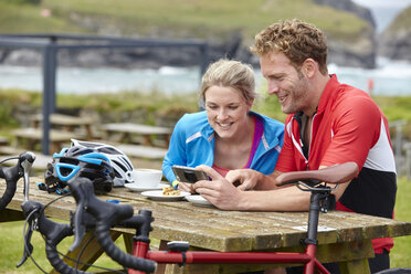 Cyclists using mobile phone at picnic table overlooking ocean - CUF22919