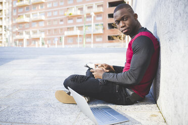 Young man sitting outdoors, leaning against wall, using digital tablet, laptop beside him - CUF22967