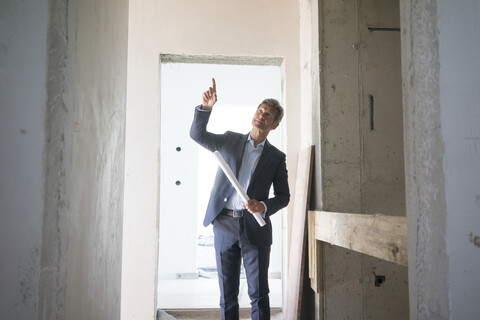 Architekt mit Bauplan, der mit dem Finger auf ein im Bau befindliches Gebäude zeigt, lizenzfreies Stockfoto