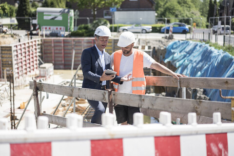 Mann im Anzug mit Tablet im Gespräch mit Bauarbeiter auf einer Baustelle, lizenzfreies Stockfoto