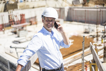 Smiling man wearing hard hat on cell phone on construction site - MOEF01305