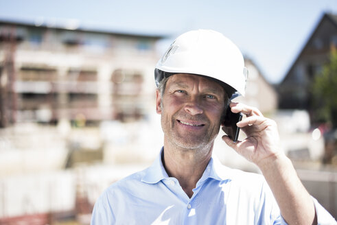 Porträt eines lächelnden Mannes mit Schutzhelm und Handy auf einer Baustelle - MOEF01307