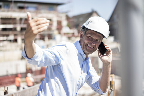 Lächelnder Mann mit Schutzhelm und Mobiltelefon auf einer Baustelle, lizenzfreies Stockfoto