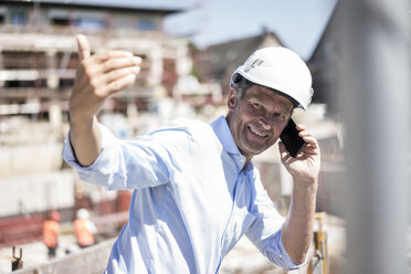 Lächelnder Mann mit Schutzhelm und Mobiltelefon auf einer Baustelle - MOEF01317