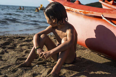 Junge sitzt am Strand bei einem Boot und schaut weg - CUF23060