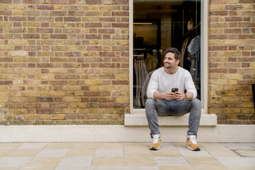 Young man with smartphone sitting on doorstep, Kings Road, London, UK - CUF23069