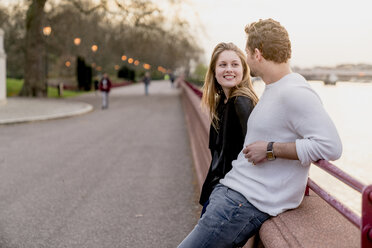 Young couple chatting on waterfront at sunset, Battersea Park, London, UK - CUF23084