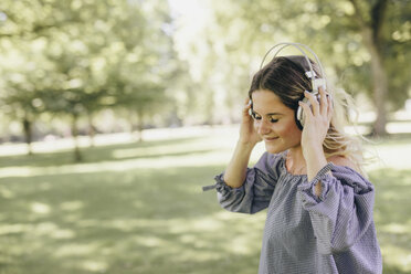 Lächelnde junge Frau in einem Park, die gerne mit Kopfhörern Musik hört - KMKF00356