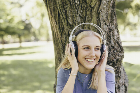 Happy young woman at tree trunk in a park wearing headphones stock photo