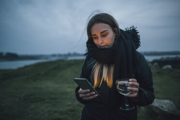 Frankreich, Bretagne, Landeda, junge Frau mit Handy und einem Glas Wein an der Küste in der Abenddämmerung - GUSF00943