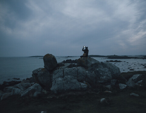 Frankreich, Bretagne, Landeda, Mann macht Handy-Foto an der Küste in der Abenddämmerung, lizenzfreies Stockfoto