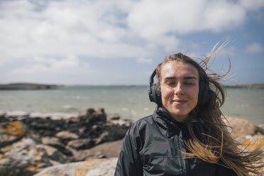 Frankreich, Bretagne, Landeda, lächelnde junge Frau mit Kopfhörern an der Küste - GUSF00955