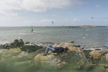 Frankreich, Bretagne, Landeda, junge Frau mit Kopfhörern auf einem Felsen an der Küste liegend - GUSF00968