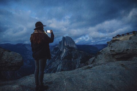 Junge Frau beim Fotografieren auf einem Felsen mit Blick auf den Yosemite-Nationalpark in der Abenddämmerung, Kalifornien, USA, lizenzfreies Stockfoto