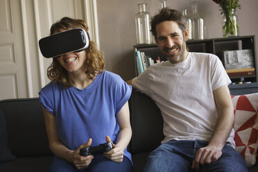 Portrait of smiling man sitting by woman playing on virtual reality headset in living room at home - FSIF03059