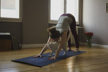 Full length of woman practicing downward facing dog position with cat on exercise mat at home - FSIF03064