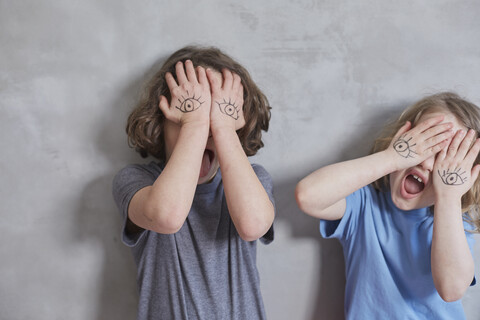 Verspielte Mädchen bedecken ihre Augen mit bemalten Händen, während sie an einer grauen Wand stehen, lizenzfreies Stockfoto