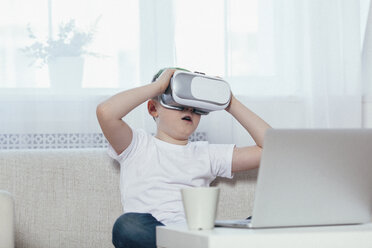 Boy wearing virtual reality headset while sitting on sofa at home - FSIF03098