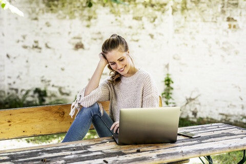 Lächelnde Frau sitzt auf einer Bank im Innenhof und schaut auf einen Laptop, lizenzfreies Stockfoto