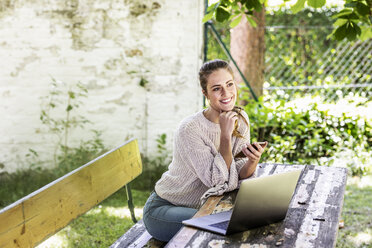 Glückliche träumende Frau im Garten sitzend mit Laptop und Handy - FMKF05082