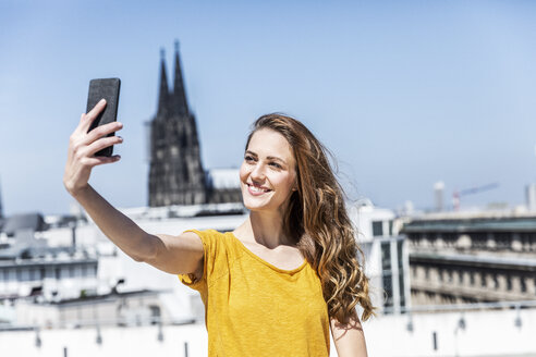 Deutschland, Köln, Porträt einer lächelnden Frau, die auf einer Dachterrasse ein Selfie mit ihrem Smartphone macht - FMKF05109