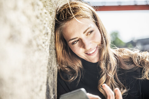 Porträt einer lächelnden Frau mit Smartphone, die an der Wand lehnt, lizenzfreies Stockfoto