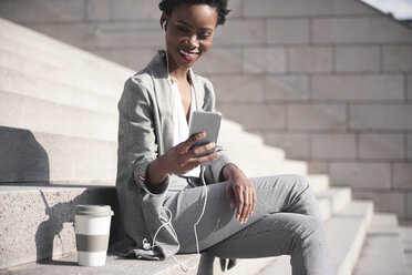 Portrait of smiling businesswoman sitting on stairs using earphones and smartphone - ABIF00542