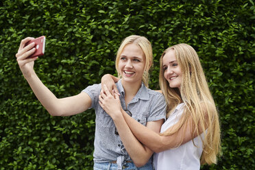 Zwei glückliche junge Frauen machen ein Selfie an einer Hecke - MMIF00144