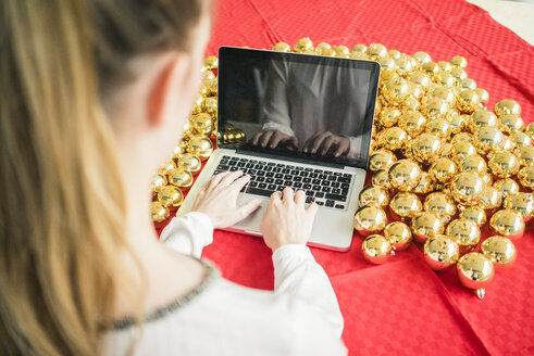 Frau sitzt am Tisch mit vielen goldenen Weihnachtskugeln und arbeitet am Laptop - MOEF01333