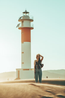 Junge Frau mit vom Wind zerzaustem Haar in Wüstenlandschaft am Leuchtturm stehend - OCAF00269