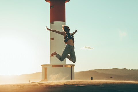 Young woman jumping in desert landscape at lighthouse stock photo