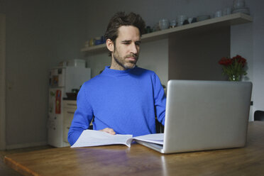 Besorgter mittlerer Erwachsener, der mit einem Laptop am Tisch in der Küche sitzt und Dokumente bearbeitet - FSIF03111