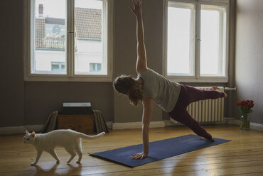 Rear view full length of woman practicing side plank pose on exercise mat by cat at home - FSIF03113