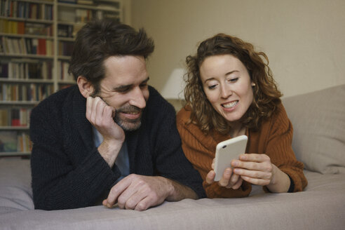 Lächelnde Frau, die ihr Smartphone mit einem Mann teilt, der zu Hause auf dem Bett liegt - FSIF03114