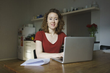Portrait of confident woman sitting with laptop and documents at table in kitchen - FSIF03117