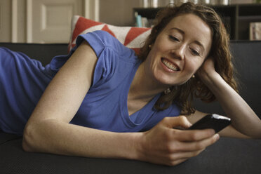 Lächelnde Frau auf dem Sofa liegend, die ihr Smartphone zu Hause benutzt - FSIF03119