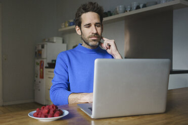 Mid adult man sitting with raspberries in plate using laptop at table in kitchen - FSIF03120